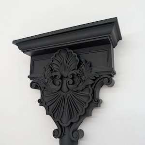 May include: A matte black wall-mounted shelf bracket featuring an ornate, carved design. The piece displays a prominent central shell motif flanked by intricate acanthus leaf scrollwork, finished with a smooth, uniform dark surface texture.