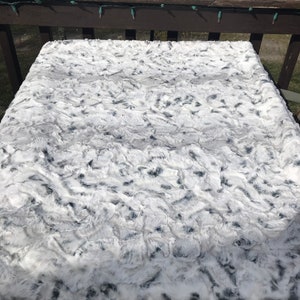 May include: A white and gray faux fur throw blanket with a wavy pattern. The blanket is lying flat on a wooden surface.