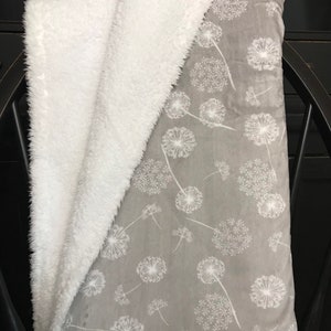 May include: A gray blanket with a white dandelion pattern and a white, fluffy sherpa lining. The blanket is folded over a black chair, showcasing the soft texture and design. The blanket is a cozy home decor item.