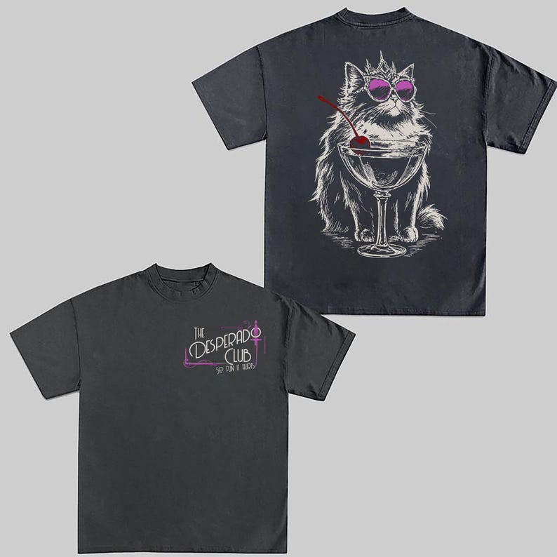 May include: Two dark gray t-shirts. One features a cat wearing sunglasses and a crown, sitting behind a cocktail glass with a cherry. The other shirt has the text "The Desperado Club" in a pink and white design.