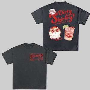 May include: Two dark gray t-shirts. One features the text "The Desperado Club" in red. The other shirt has a graphic of a cocktail and a cat wearing sunglasses, with the text "Dirty Shirley" and "Mama needs a night off."