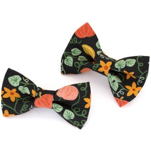 May include: Two black bow ties with a repeating pattern of orange pumpkins, yellow flowers, and green leaves. The bow ties are made of fabric and are set against a white background. The bow ties are likely for pets or children.