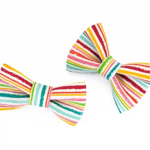 May include: Two colorful bow ties with a striped pattern in shades of red, yellow, green, blue, and pink. The bow ties are made of fabric and have a white background.