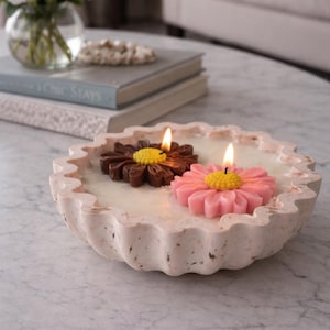May include: A decorative, scalloped-edge bowl holds two lit flower-shaped candles. One candle is brown with a yellow center, the other is pink with a yellow center. The bowl is light pink and sits on a marble surface.