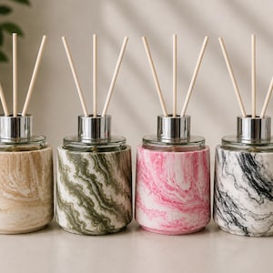 May include: Four cylindrical reed diffusers with marble-patterned bases in beige, green, pink, and black. Each glass vessel features a silver-toned metal collar and three light-colored wooden reeds extending upward from the center of the container.