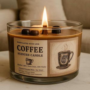 May include: A brown glass candle with a coffee scent. The candle has a label that says "MARRCLAFTED WITH LOVE COFFEE SCENTED CANDLE" and features a drawing of a cup of coffee. The candle is lit and has a flame.