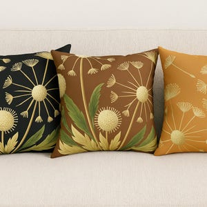 May include: Three square throw pillows with dandelion designs. One pillow is black with a gold dandelion, another is brown with a gold dandelion and green leaves, and the third is orange with a gold dandelion.