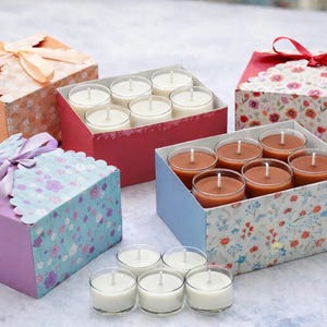 May include: Several decorative gift boxes with candles. The boxes are in various colors, including orange, pink, and purple, and are decorated with floral patterns and ribbons. The candles are in clear glass holders.