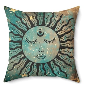 May include: A teal and gold throw pillow with a black and white illustration of a sun with a face and closed eyes. The sun has a crescent moon and three dots above its head. The background is a teal and gold galaxy with stars.