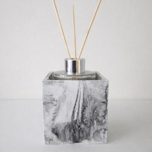 May include: A decorative reed diffuser with a marbled gray and white square base. The glass bottle has a silver collar and holds wooden reeds for fragrance. The design is minimalist and modern.