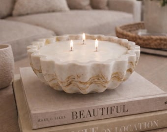 May include: A three-wick candle in a round, scalloped-edge white and gold marble-patterned bowl. The wax is white and the three wicks are lit, producing small flames. The bowl has a wavy, textured exterior with gold veining throughout.