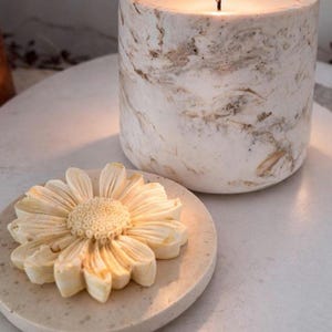May include: A lit candle in a marble-patterned cylindrical container, next to a decorative flower-shaped coaster. The candle's wax is white, and the container has brown and white marbling. The coaster is beige, with a detailed flower design.