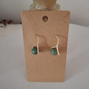 May include: A pair of gold earrings with green gemstone accents. The earrings are displayed on a brown card.