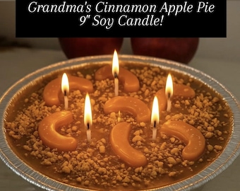 May include: A 9-inch soy candle designed to resemble a cinnamon apple pie. The candle features orange wax 'apple slices' and a crumbly topping, with lit wicks mimicking flames. The text reads "Grandma's Cinnamon Apple Pie 9" Soy Candle!"