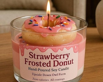 Peut inclure: Une bougie en forme de donut glacé à la fraise avec des vermicelles colorés, surmontée d'une mèche allumée. Le pot en verre porte l'inscription "Strawberry Frosted Donut".