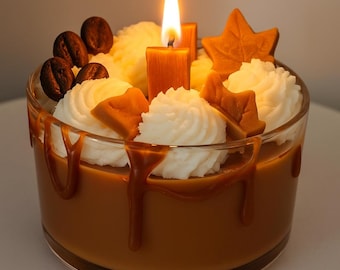 May include: A candle in a clear glass container, resembling a dessert. The candle has a caramel-colored wax base with white whipped cream, coffee beans, and maple leaf-shaped decorations. A lit candle sits in the center, with caramel drips down the side.