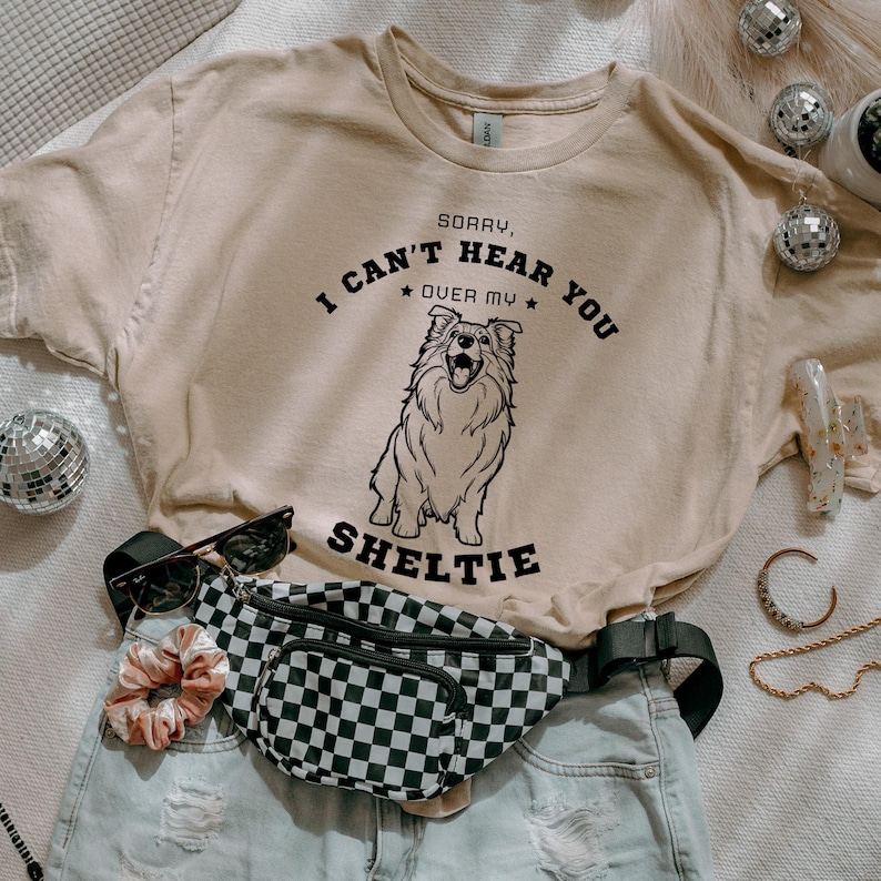 Peut inclure: Un t-shirt beige avec un motif noir qui dit "D&eacute;sol&eacute;, je n'entends pas au-dessus de mon Sheltie" avec une illustration de chien Sheltie de dessin anim&eacute;. Le t-shirt est pos&eacute; sur une surface blanche avec une banane noire et blanche &agrave; carreaux, une paire de lunettes de soleil et un chouchou.