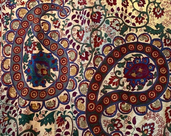 May include: A white fabric with a paisley pattern embroidered in shades of red, blue, green, and gold. The fabric is decorated with intricate floral designs and paisley motifs.