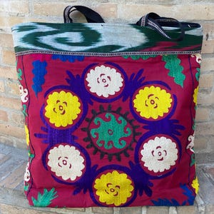 Può includere: Grande borsa tote con un vivace design ricamato. La borsa presenta uno sfondo rosso intenso con motivi floreali gialli e bianchi ricamati e una parte superiore a fantasia verde e blu. I manici sono neri.