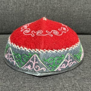 Può includere: Un berretto ricamato rosso, grigio e verde con la parte superiore arrotondata. Il berretto presenta cuciture bianche e un motivo decorativo attorno alla base. Il berretto ha un bordo color argento.