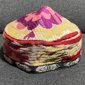 Puede incluir: Un gorro en forma de cúpula con intrincados bordados en tonos magenta, rosa, amarillo y rojo sobre una base de tela beige. El borde inferior está rematado con una banda negra decorada con un patrón geométrico blanco y costuras horizontales.