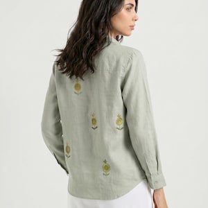 Puede incluir: Una camisa de manga larga en color verde salvia con botones y un patrón disperso de pequeños motivos florales bordados en amarillo y verde. La camisa cuenta con cuello clásico, canesú trasero recto y dobladillo redondeado, combinada con pantalones blancos.