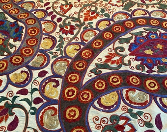 May include: A white fabric with intricate embroidery in shades of blue, red, green, and gold. The design features paisley patterns, floral motifs, and circular shapes.