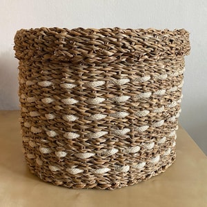 Puede incluir: Una cesta redonda tejida de color marrón natural con detalles blancos. La cesta está hecha de materiales naturales y tiene un diseño decorativo.