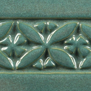 May include: A close-up of a teal green ceramic tile with a raised embossed pattern of repeating four-leaf clover designs.