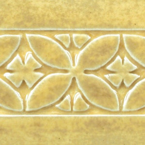 May include: A close-up of a yellow ceramic tile with a raised, white, floral design. The tile has a textured surface and a subtle sheen.