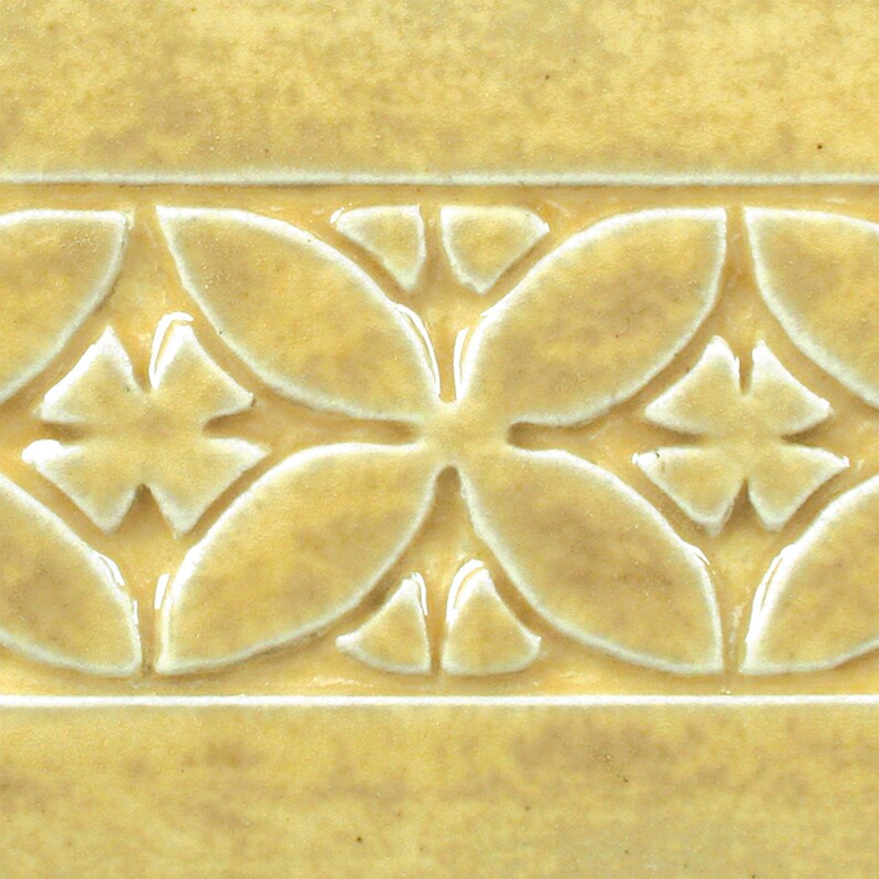 May include: A close-up of a yellow ceramic tile with a raised, white, floral design. The tile has a textured surface and a subtle sheen.