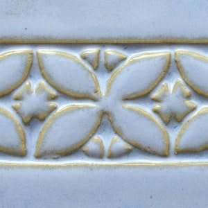 May include: A close-up of a light blue ceramic tile with a raised pattern of gold flowers. The tile has a smooth, glossy finish.