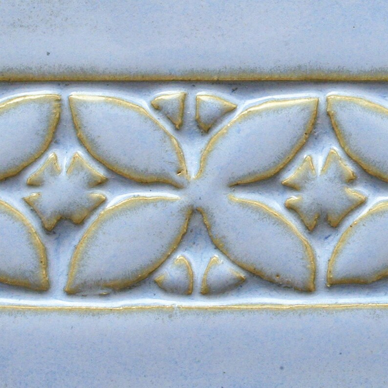 May include: A close-up of a light blue ceramic tile with a raised pattern of gold flowers. The tile has a smooth, glossy finish.