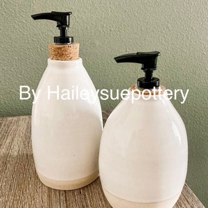 May include: Two white ceramic soap dispensers with black pump tops and cork stoppers. The dispensers have a rounded shape with a natural beige base. The text "By Haileysuepottery" is visible.