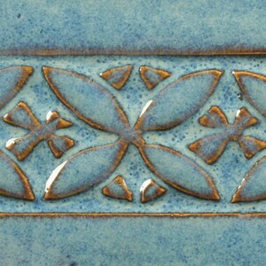 May include: A close-up of a blue ceramic tile with a brown embossed pattern. The pattern is a repeating design of four-leaf clover shapes.