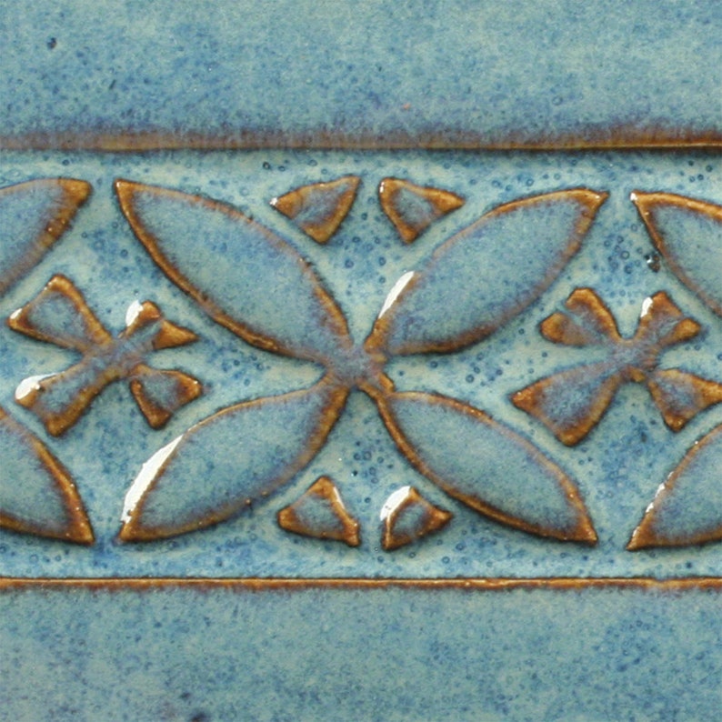 May include: A close-up of a blue ceramic tile with a brown embossed pattern. The pattern is a repeating design of four-leaf clover shapes.