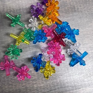 May include: Assortment of colorful plastic Christmas tree ornaments in various shapes, including stars, flowers, and snowflakes. The ornaments are scattered on a gray wooden surface.