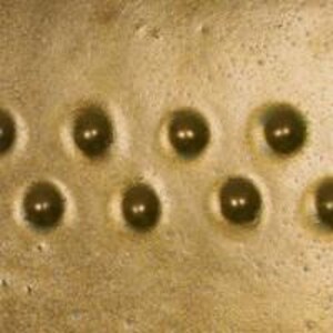 Pu&ograve; includere: Primo piano di una superficie metallica dorata con nove rientranze circolari equidistanti. Ogni rientranza contiene una piccola perla metallica rotonda.