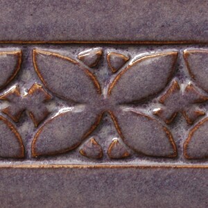 May include: A close-up of a purple ceramic tile with a raised embossed design of a repeating floral pattern in a copper brown glaze.