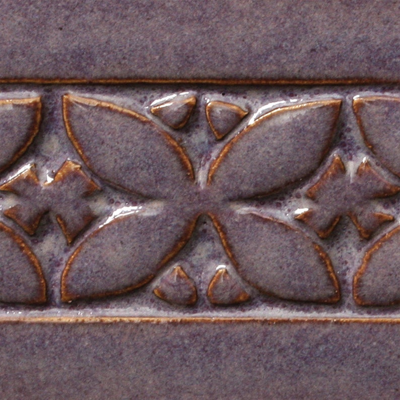 May include: A close-up of a purple ceramic tile with a raised embossed design of a repeating floral pattern in a copper brown glaze.