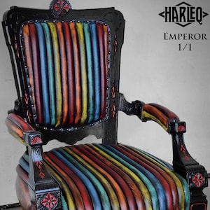 Op de afbeelding: Antieke fauteuil met een zwart frame en een zitting en rugleuning bekleed met verticale strepen in levendige kleuren. De stoel heeft sierlijke details in rood en zwart, met kleine wieltjes op de poten.