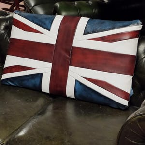 May include: Rectangular cushion displaying the Union Jack flag. The design features a blue backdrop with white and red stripes. The cushion is placed on a dark green leather sofa.