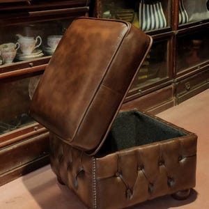May include: A brown leather ottoman with a tufted top and a hinged lid. The ottoman is open, revealing a black fabric lining. The ottoman is sitting on a wooden floor.