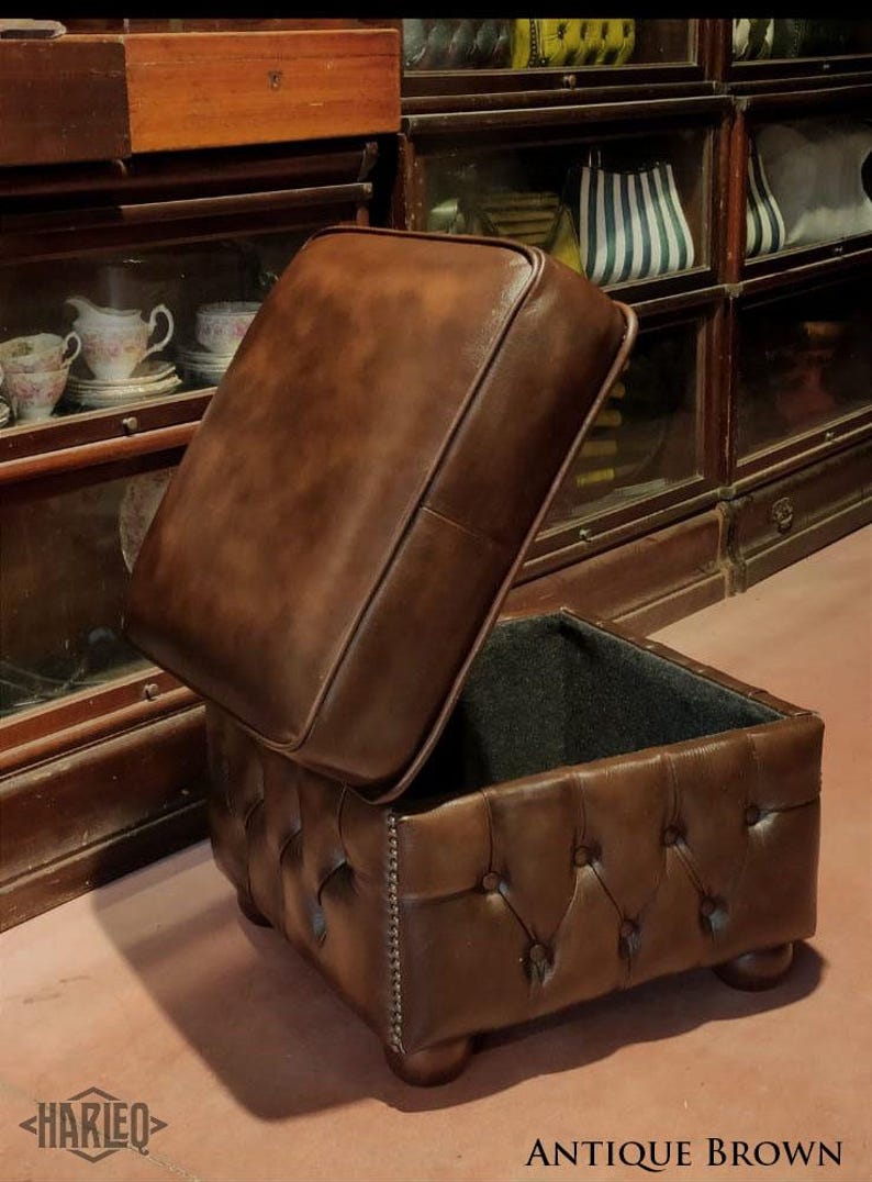 May include: A brown leather ottoman with a tufted top and a hinged lid. The ottoman is open, revealing a black fabric lining. The ottoman is sitting on a wooden floor.