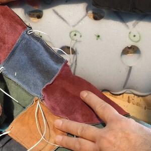 May include: Close-up of a patchwork design in progress. Various colored suede squares, including blue, burgundy, and brown, are stitched together with white thread. The work is being done on a light-colored foam base.