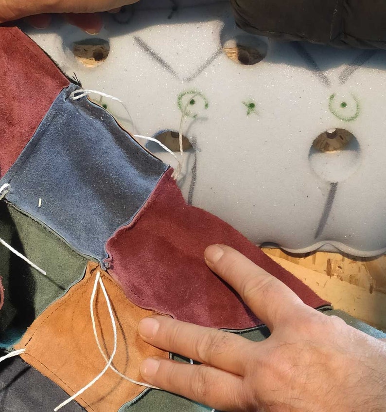 May include: Close-up of a patchwork design in progress. Various colored suede squares, including blue, burgundy, and brown, are stitched together with white thread. The work is being done on a light-colored foam base.