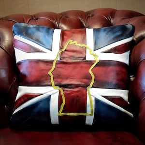 May include: A cushion with a Union Jack design in red, white, and navy blue leather. A gold outline of a shape is centered on the cushion. The cushion is placed on a red leather armchair.
