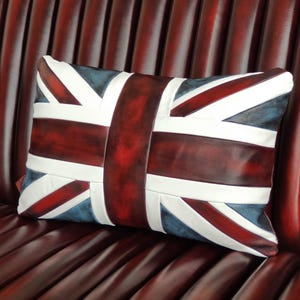 Pu&ograve; includere: Cuscino rettangolare con il design della bandiera Union Jack. Il cuscino ha una combinazione di colori rosso scuro, bianco e blu. Il cuscino &egrave; posto su una superficie in pelle rosso scuro.