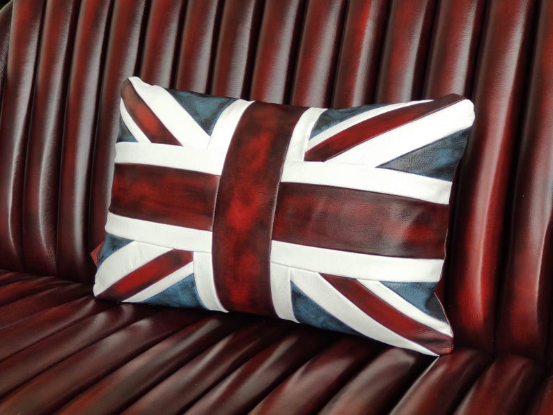 Pu&ograve; includere: Cuscino rettangolare con il design della bandiera Union Jack. Il cuscino ha una combinazione di colori rosso scuro, bianco e blu. Il cuscino &egrave; posto su una superficie in pelle rosso scuro.