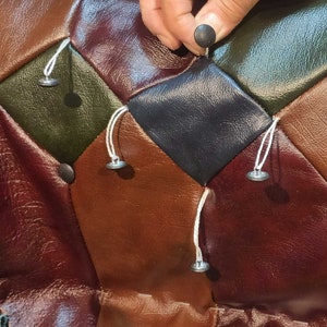 May include: Close-up of a multi-colored, diamond-patterned leather surface. The colors include shades of brown, green, and black. A hand is shown placing a black button. Silver buttons are attached with white string.